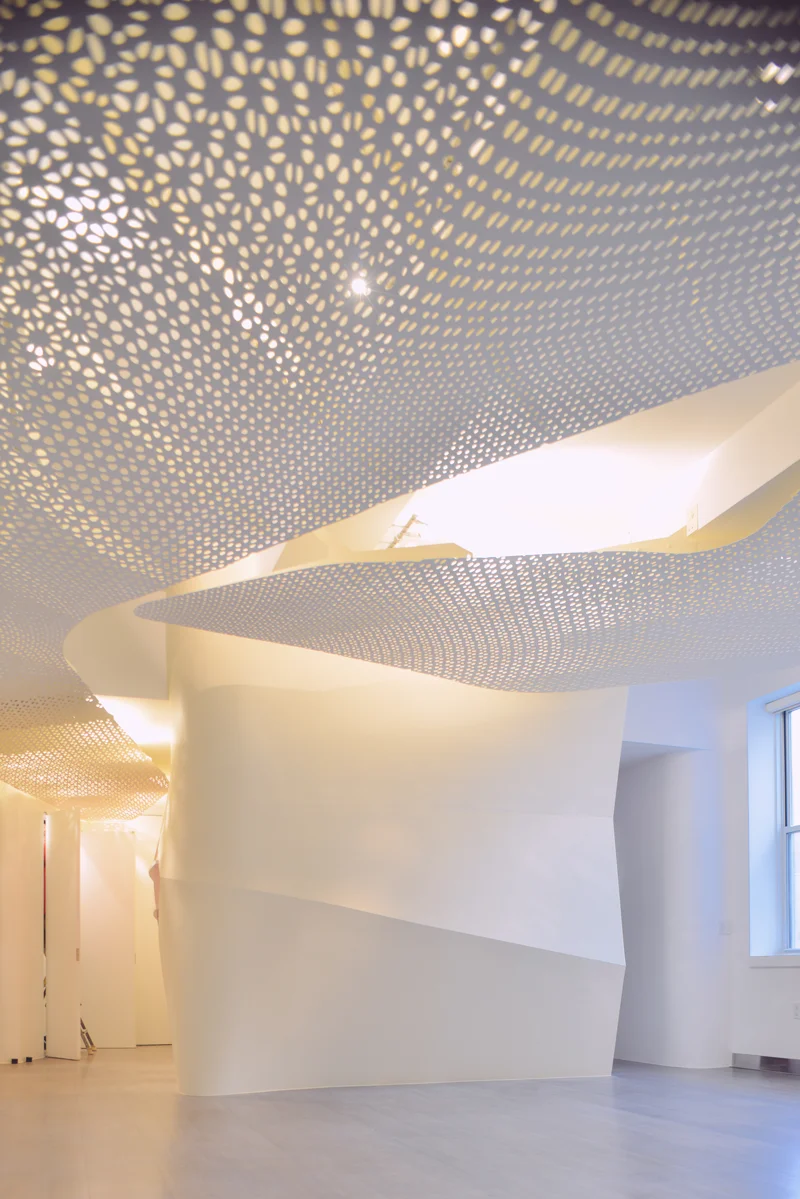 A white perforated ceiling surface curving through a gallery interior, daylight passing through thousands of petal-shaped openings.
